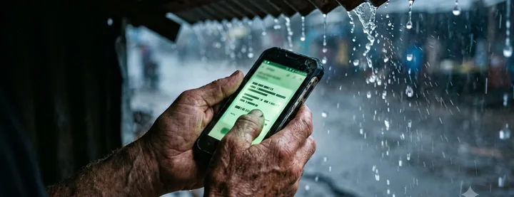 Weathered hands holding a phone showing a mobile-money payout confirmation as rain falls